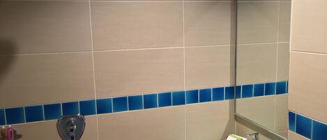 A bathroom with beige tiles, a blue-tiled accent strip, a glass sink, a handheld showerhead, and a mirror.