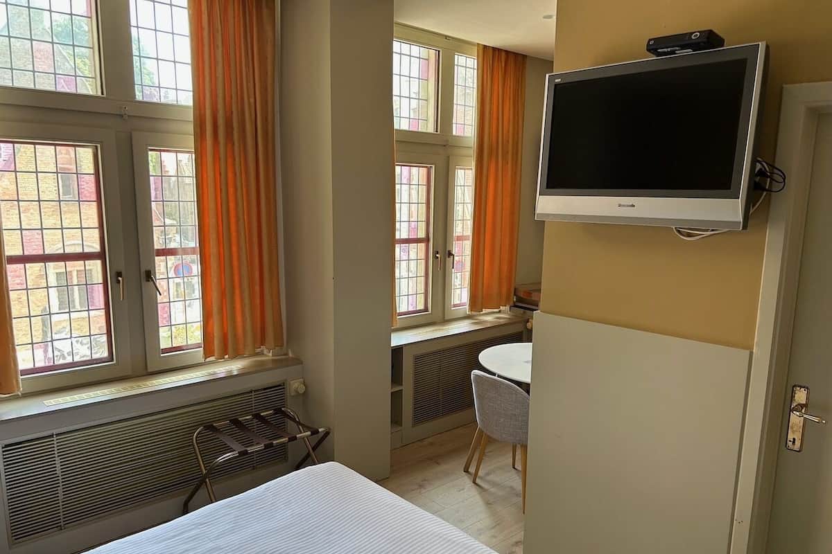 Quarto de hotel com cama, televisão na parede, mesa pequena com cadeira e grandes janelas com cortinas.