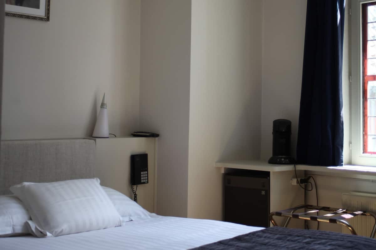 Quarto de hotel com cama, mesa de cabeceira, abajur, cafeteira e janela com cortinas.