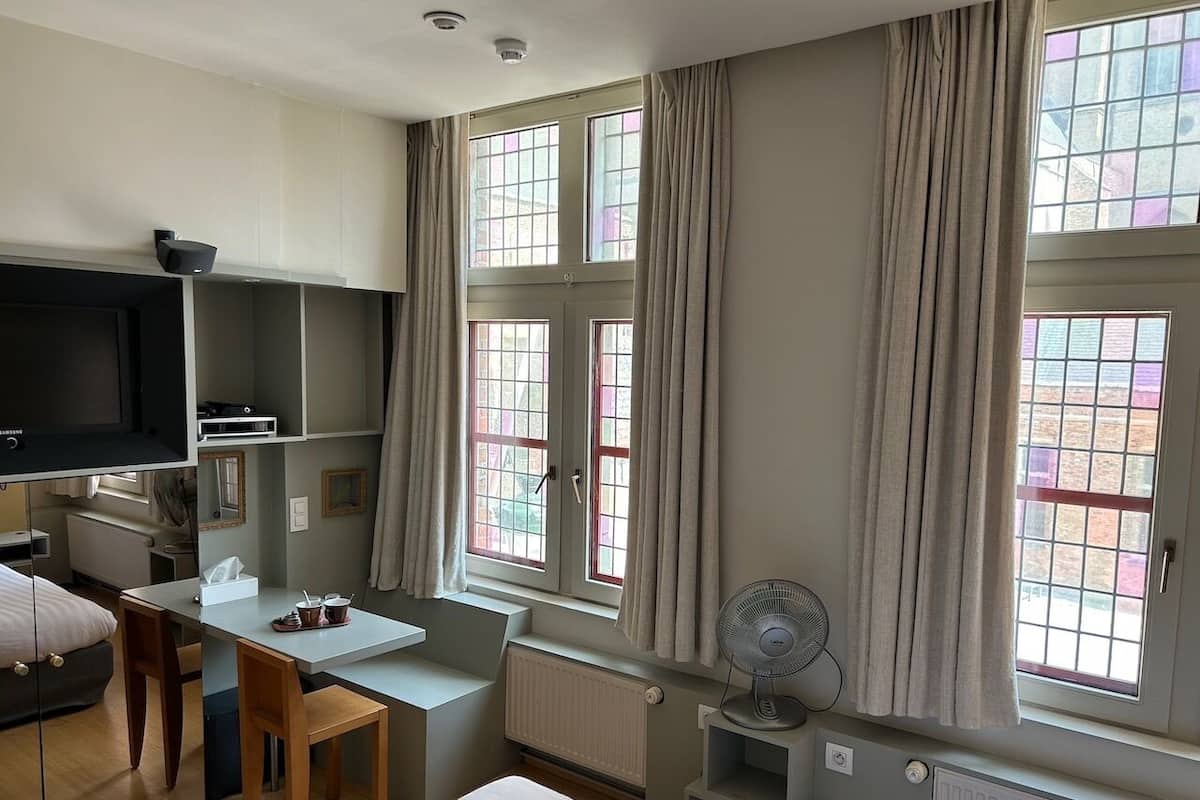 Quarto de hotel com cama, escrivaninha, cadeira, mesa, ventoinha e janelas amplas com cortinas.