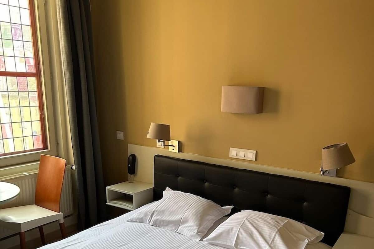 Quarto de hotel com uma cama grande, uma cadeira, uma mesa de cabeceira e uma janela com cortinas.