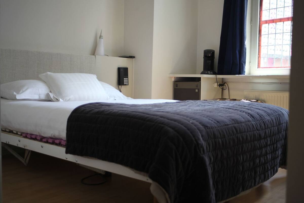 Uma cama de solteiro com cabeceira branca, mesa de cabeceira, abajur, telefone e janela com cortinas.