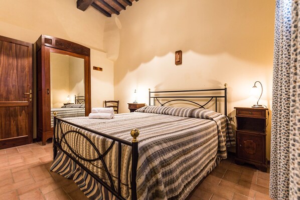 A bedroom with a bed, a wooden wardrobe, a mirror, a nightstand, and a lamp.