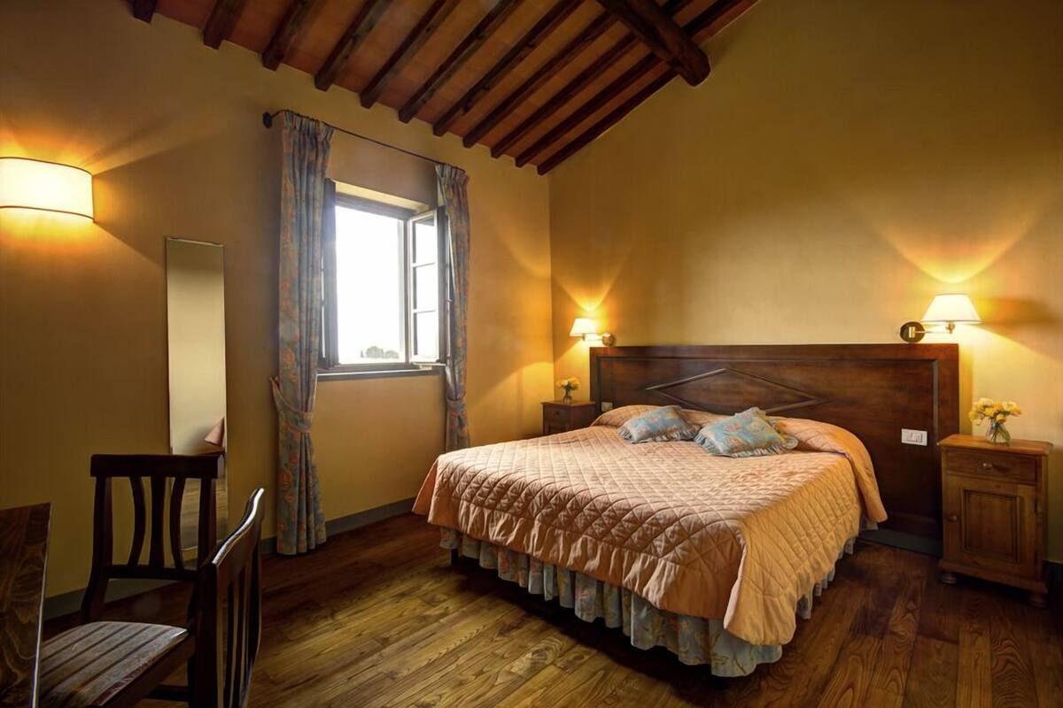 Een slaapkamer met een houten bed, twee bedlampen, een raam met gordijnen, een houten stoel en een klein kastje.