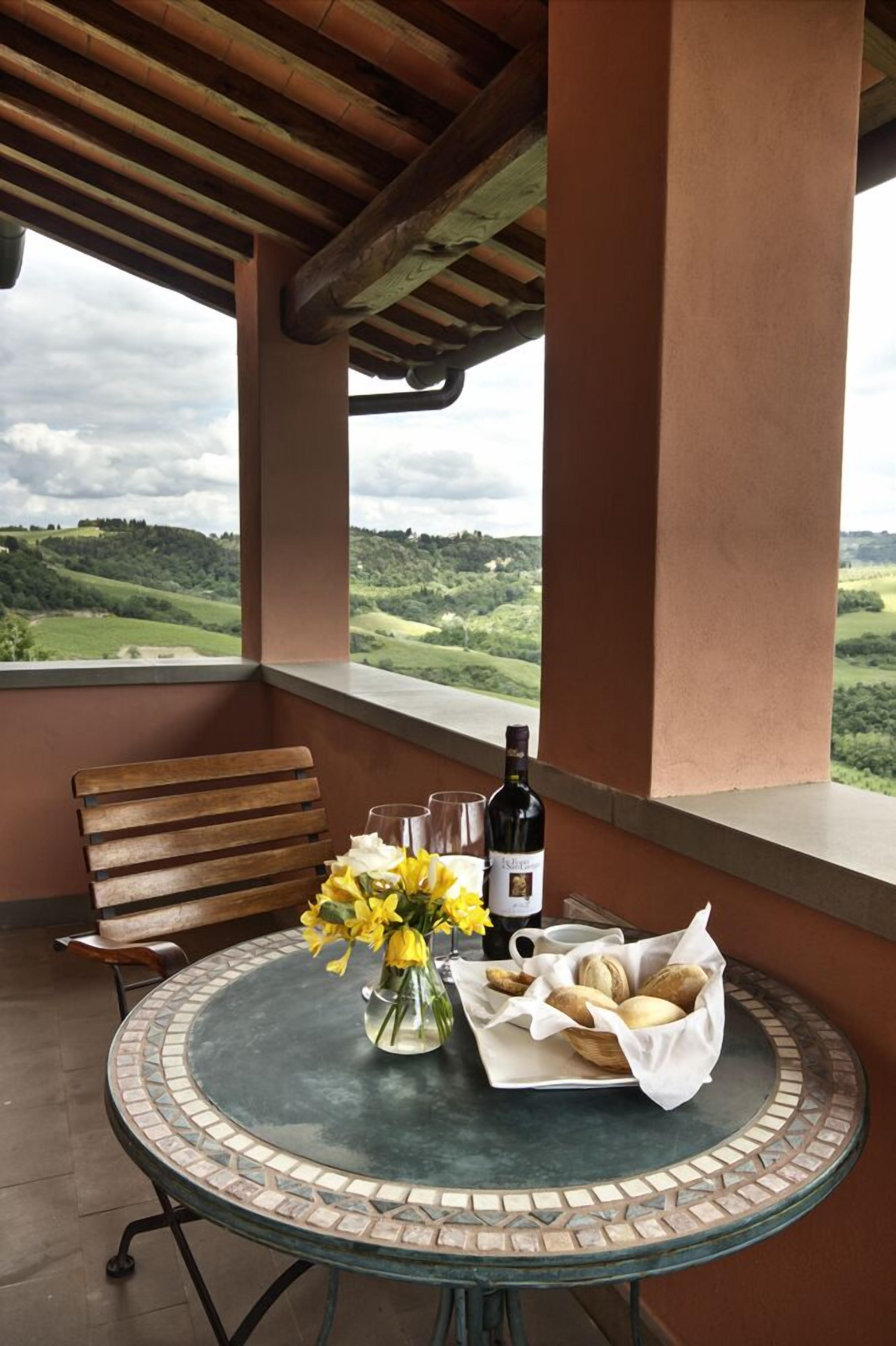 Une table ronde avec une bouteille de vin, des verres, un vase rempli de fleurs jaunes et un panier de pain, le tout sur un balcon offrant une vue sur des collines verdoyantes.
