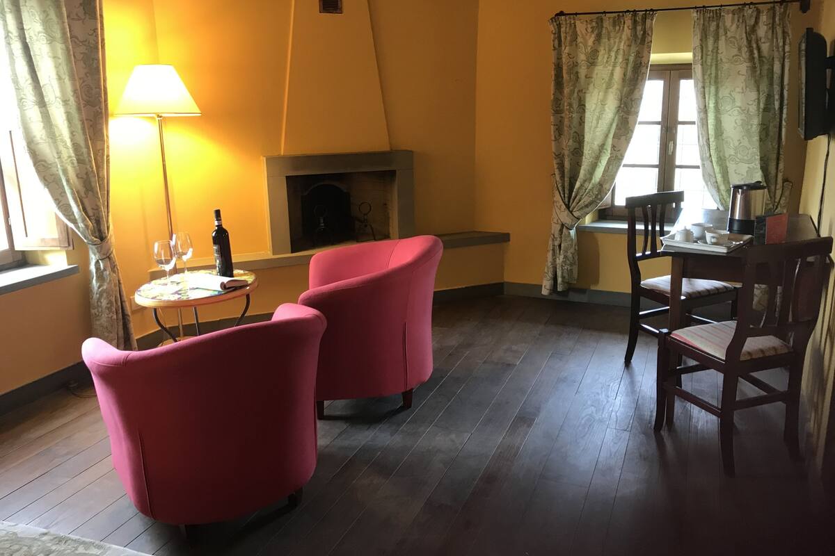 Een gezellige kamer met een open haard, twee rode fauteuils, een eettafel met stoelen en een lamp.