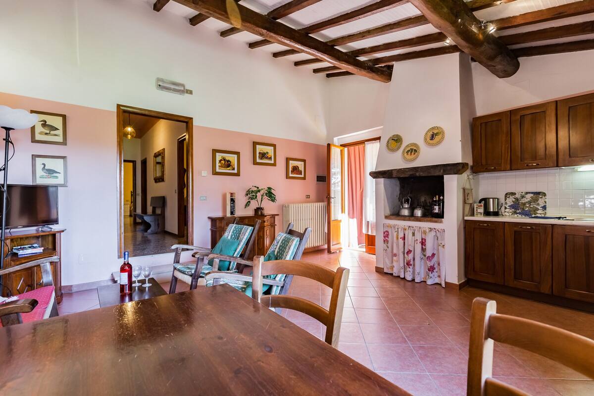 Een eetgedeelte met een houten tafel en stoelen, een keuken met houten kastjes, en een woonkamer met een televisie en ingekaderde foto's aan de muur.