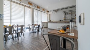 A modern dining area with wooden tables and chairs, a kitchen area with shelves, and large windows with blinds. - Villa Rocla Guest House Pompei (Pompei)