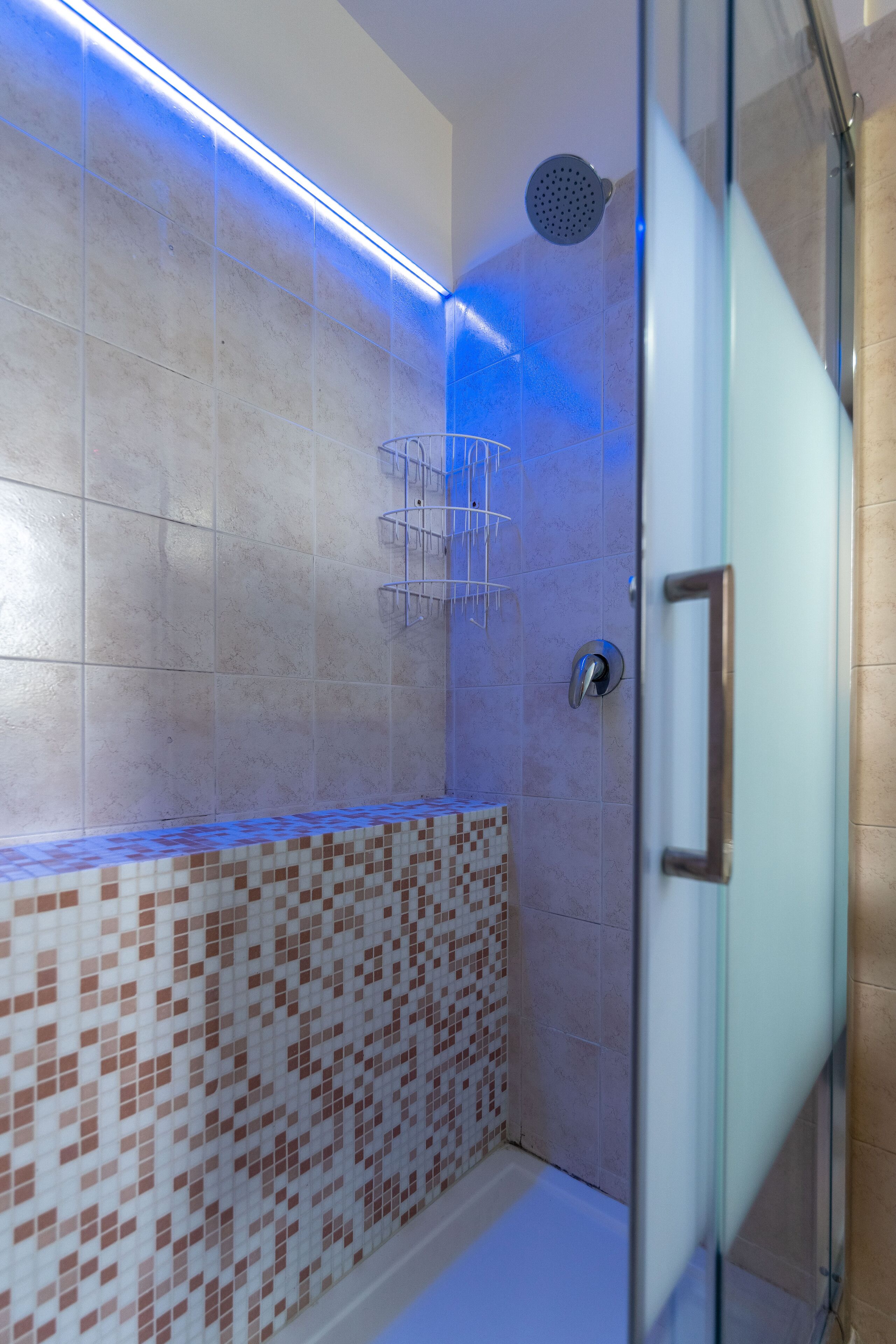 A shower with a glass door, a tiled wall with a blue light strip, and a showerhead.