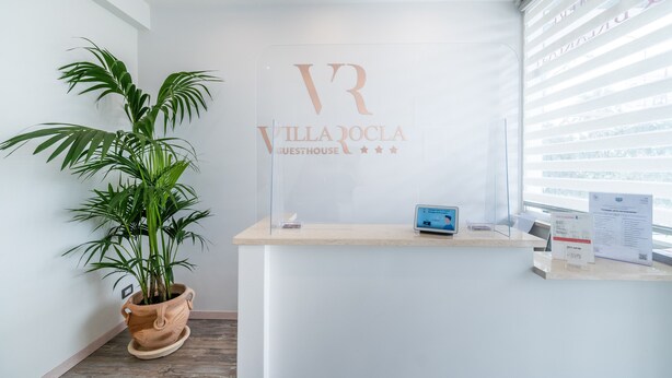 A reception desk with a potted plant, a sign reading "Villa Rocla Guesthouse", and a window with blinds. - Villa Rocla Guest House Pompei (Pompei)