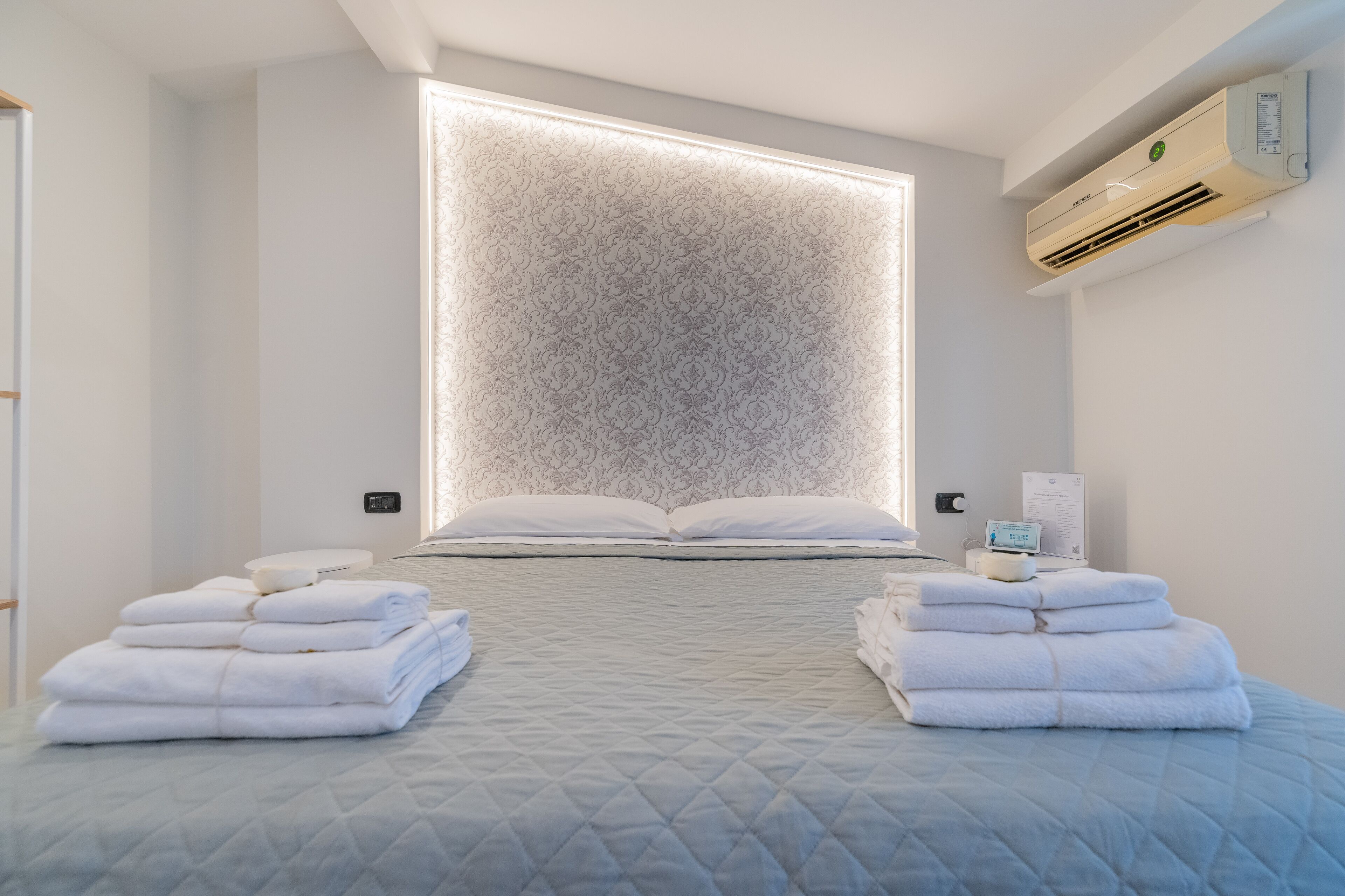 A modern hotel room with a large bed, a wall-mounted air conditioner, and neatly folded towels on either side of the bed.