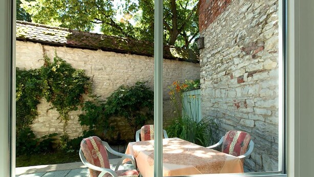 A glass-enclosed patio with a table and chairs, a stone wall, and greenery. - Hotel am Markt (Greding)