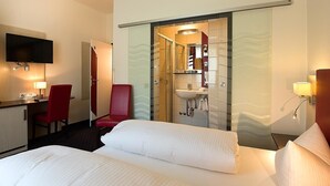 A hotel room with a bed, a desk with a TV, a red chair, and a bathroom with a mirror and sink. - Hotel am Markt (Greding)