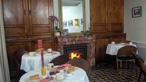 Un comedor con dos mesas redondas, cada una con platos, tazas y un pequeño jarrón con flores. Una chimenea con fuego visible se encuentra entre las mesas.