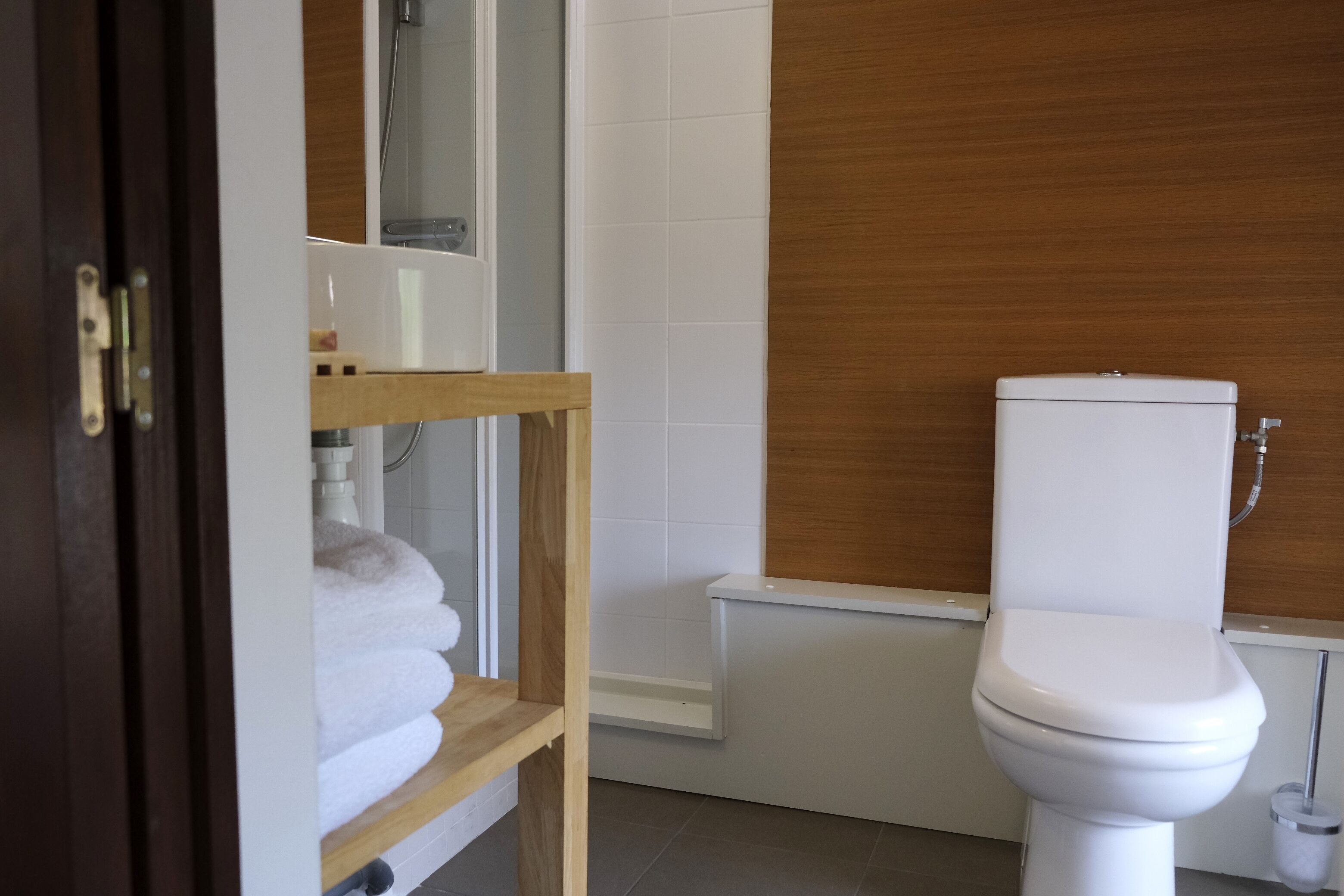 A bathroom with a white toilet, a wooden shelf with towels, and a white tiled wall.