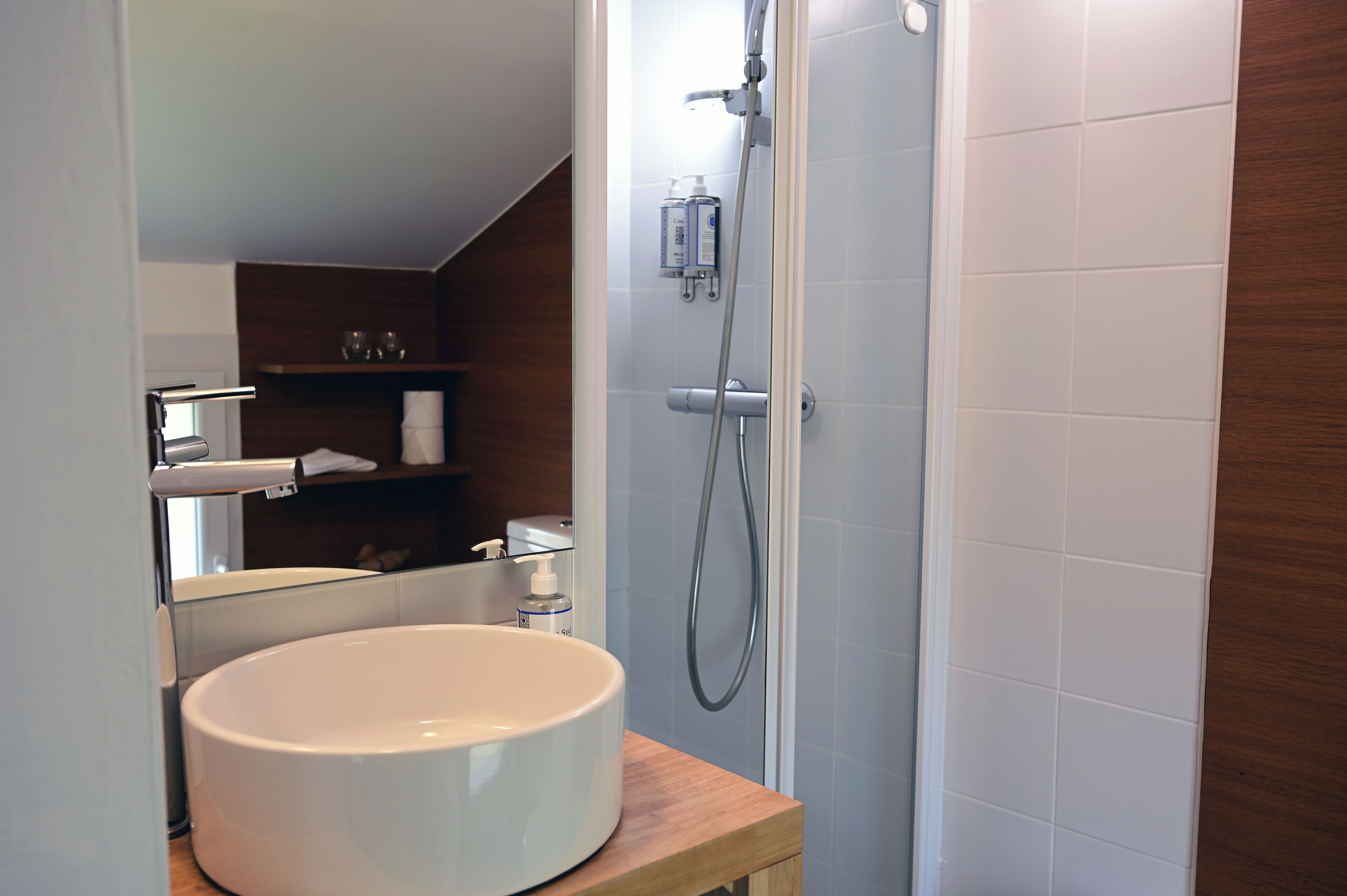 A bathroom with a white sink, a mirror, a shower, and a wooden shelf with toiletries.