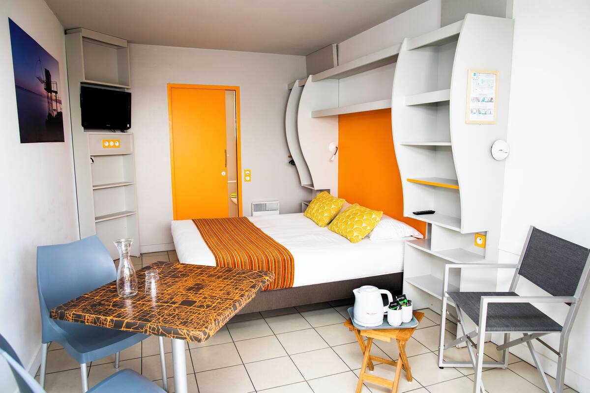 Ein Hotelzimmer mit Bett, Fernseher, Tisch mit Stühlen, einem kleinen Tisch mit Wasserkocher und einem Sessel.