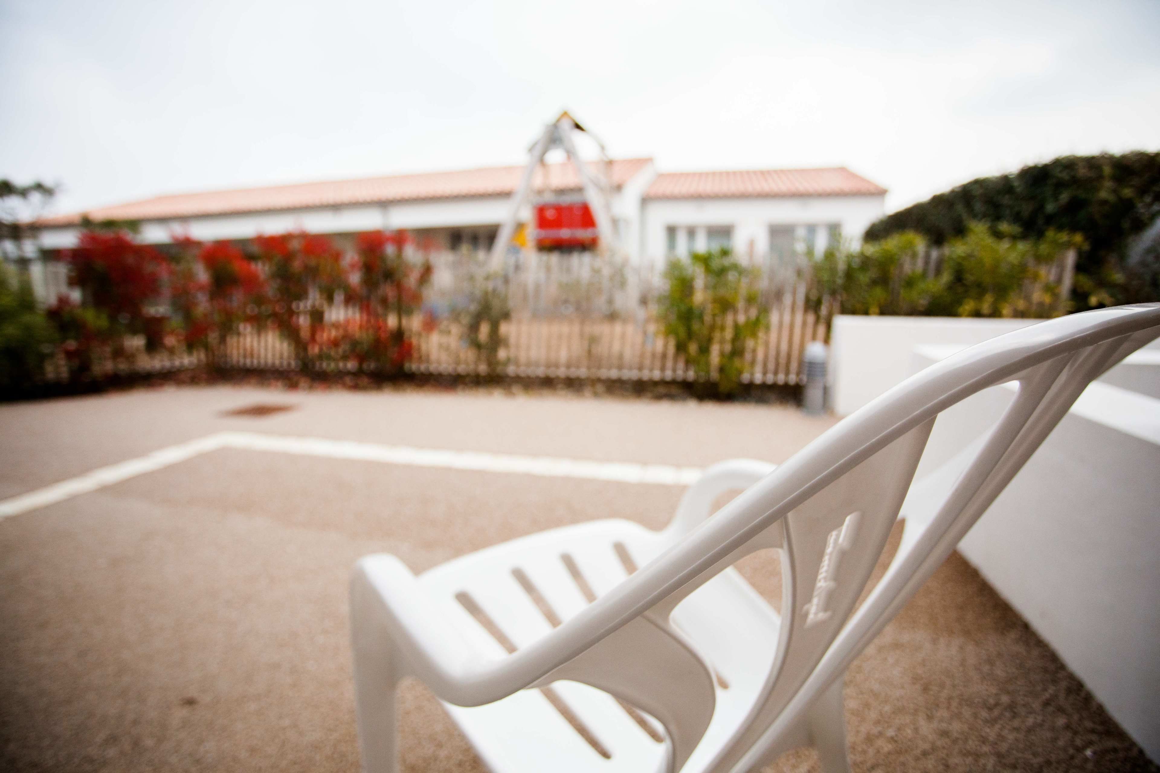 Au premier plan, une chaise en plastique blanche, avec un arrière-plan flou où l’on aperçoit un bâtiment avec une enseigne rouge et une clôture ornée de fleurs rouges.