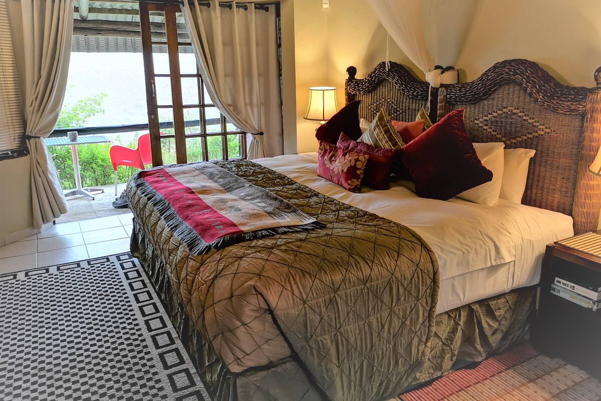 Ein Schlafzimmer mit Bett, Kissen, Nachttisch, Lampe und Blick durch das Fenster.