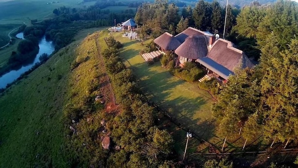 An aerial view of a property with a river, a thatched-roof building, and a well-maintained lawn. - Amazian Mountain River Lodge (Underberg)
