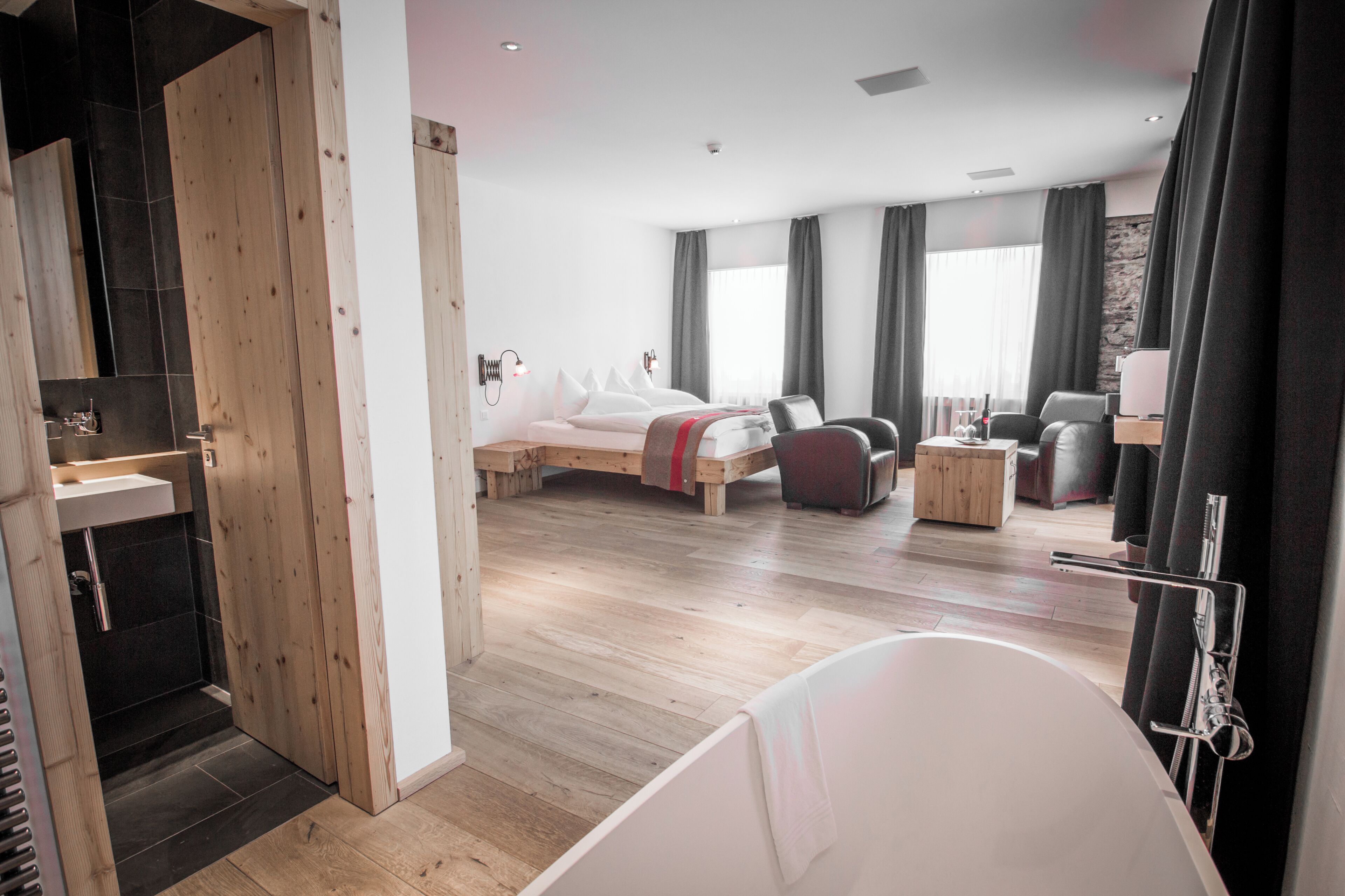 Una habitación de hotel moderna con piso de madera, una cama grande, una zona de estar con sofá y sillones, una mesita y una bañera.