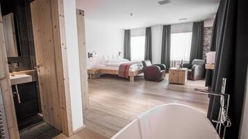 Una habitación de hotel moderna con piso de madera, una cama grande, una zona de estar con sofá y sillones, una mesita y una bañera.