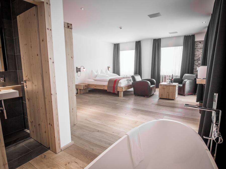 Una habitación de hotel moderna con piso de madera, una cama grande, una zona de estar con sofá y sillones, una mesita y una bañera.