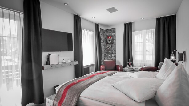Une chambre d’hôtel moderne avec un grand lit, un téléviseur fixé au mur, un mur décoratif en pierre et un plaid rouge et blanc à rayures.