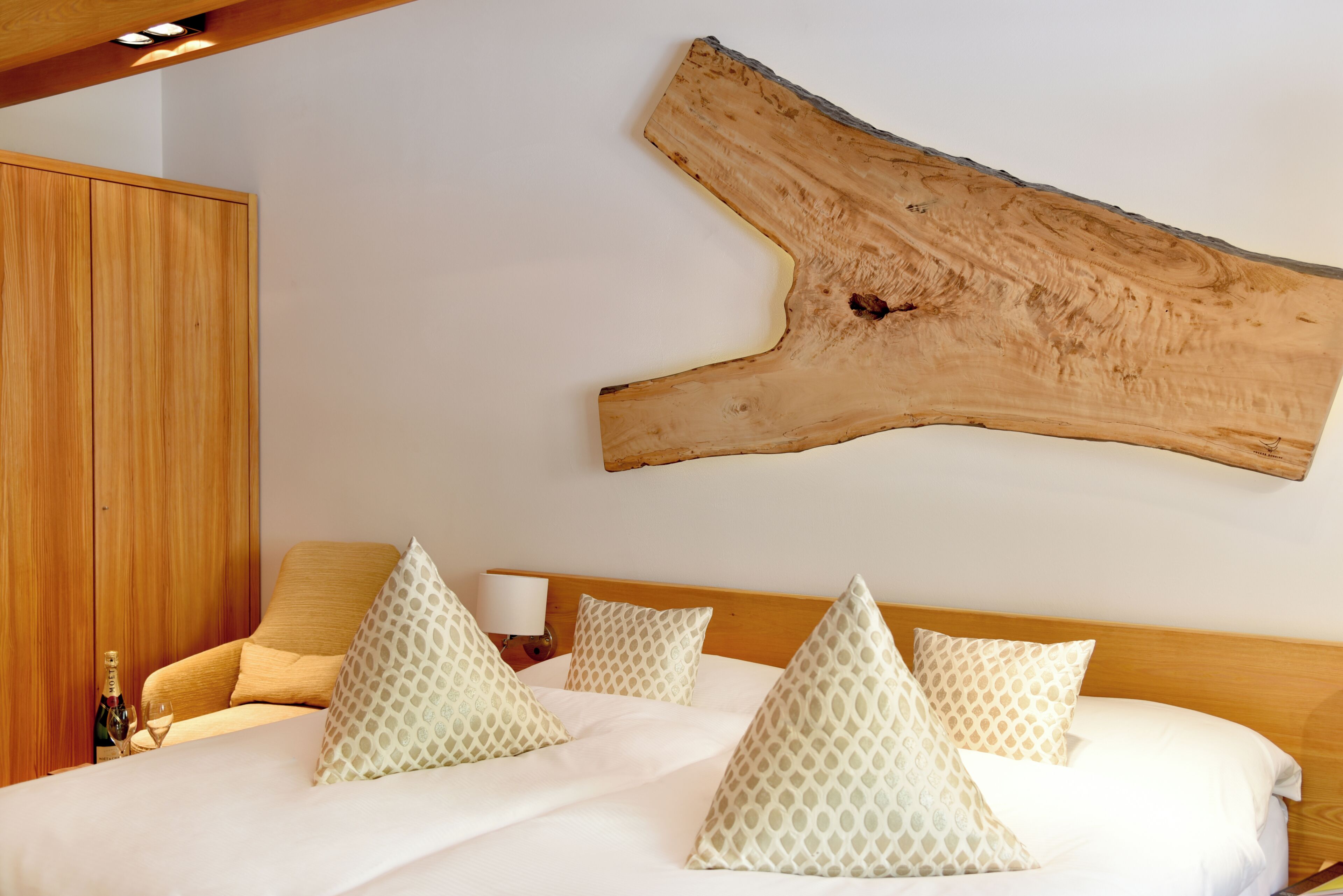 Une chambre avec une grande poutre en bois au mur, un lit avec une literie blanche et des coussins à motifs.