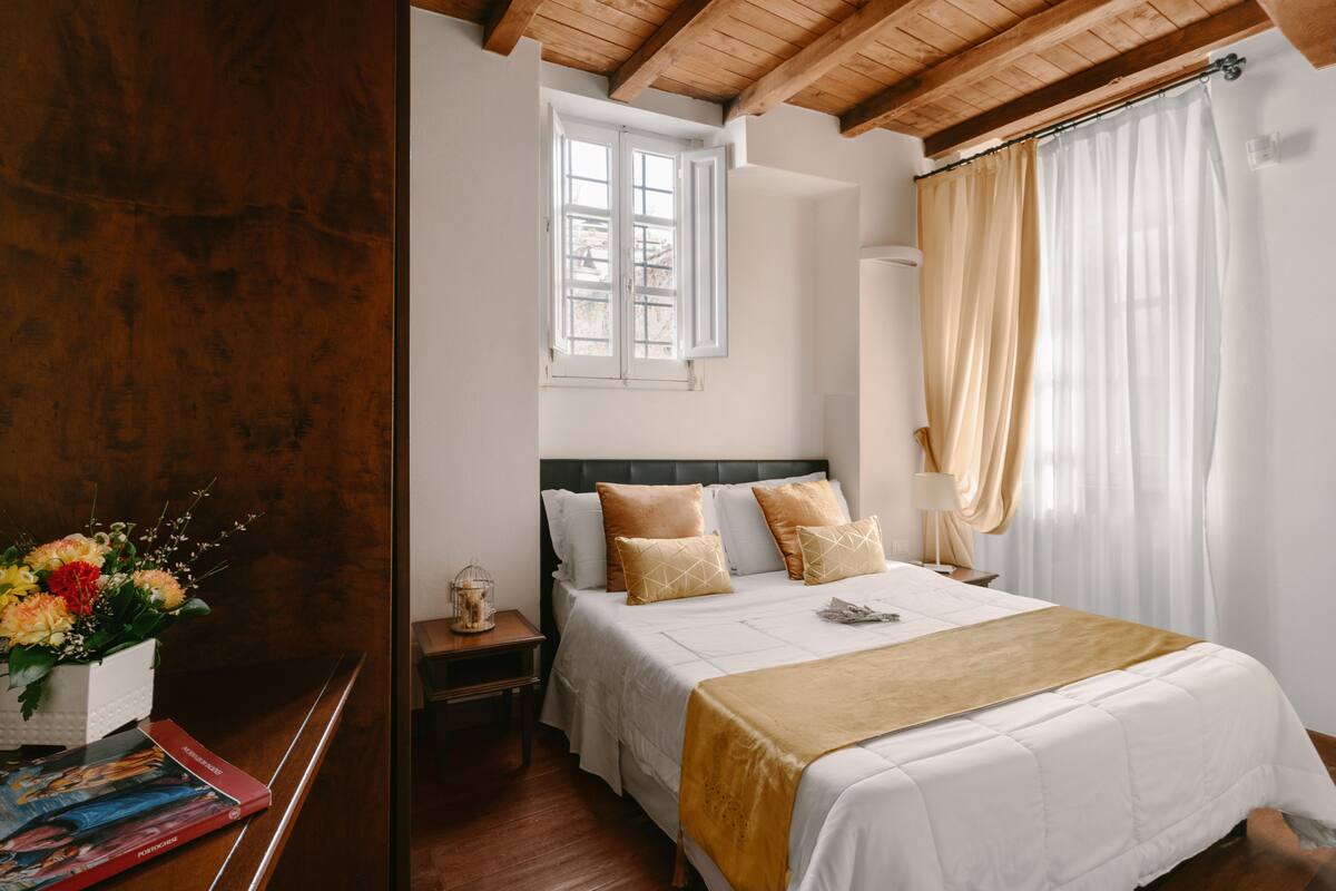 A bedroom with a wooden ceiling, a large bed with white and beige bedding, a nightstand with a lamp, a window with curtains, and a vase with flowers on a wooden table.