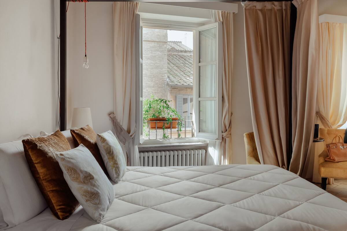 A bedroom with a large bed, a window with curtains, a radiator, and a chair.