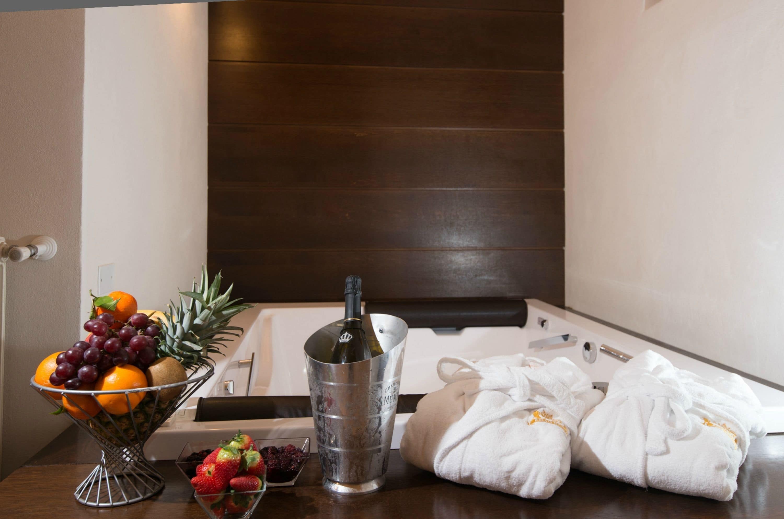 A hotel room with a wooden headboard, a fruit bowl, a bottle of wine, and towels on the table.