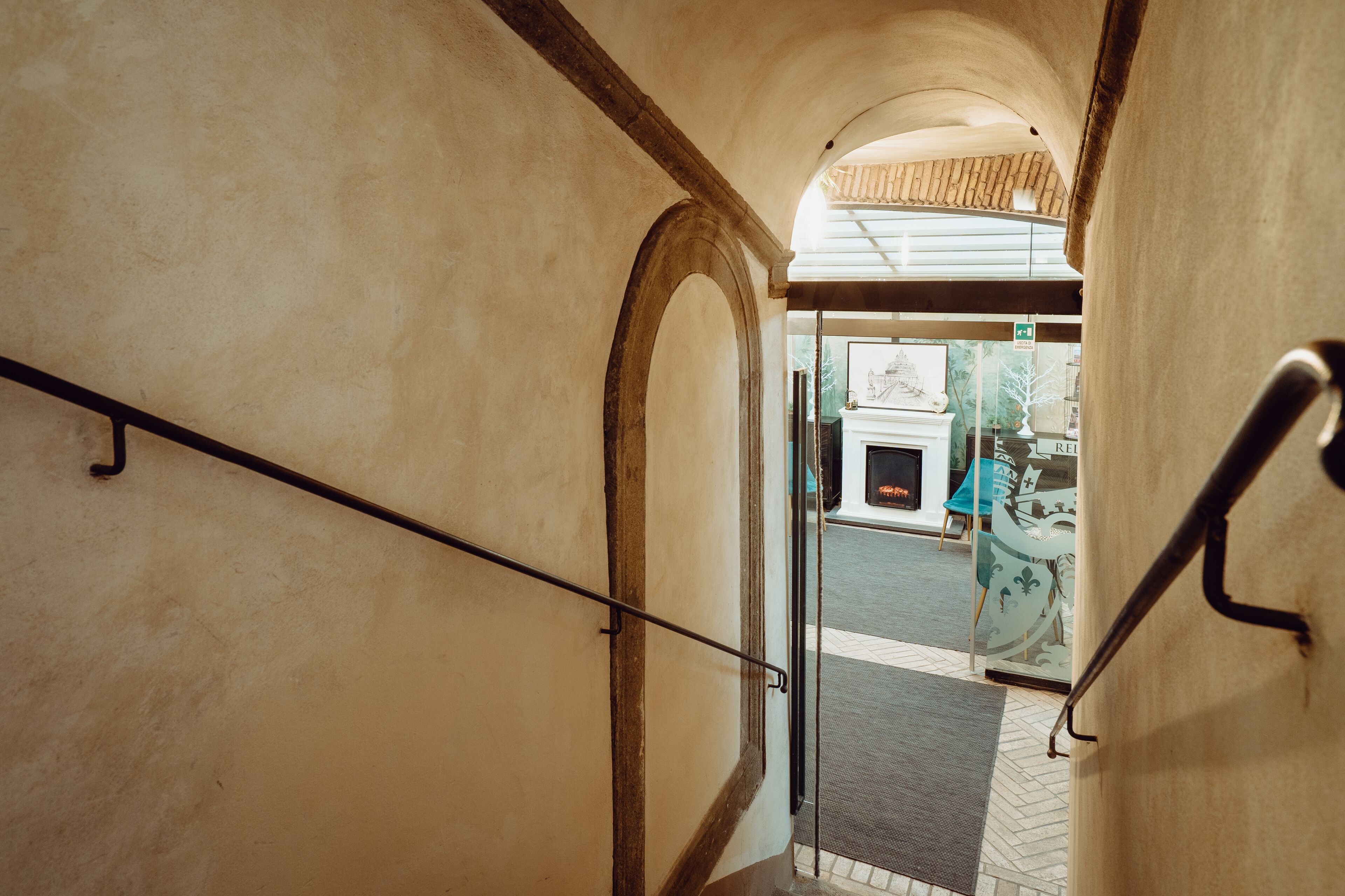 A hallway with a metal handrail, an arched doorway, and a tiled floor leading to an outdoor area with a fire extinguisher and a sign.