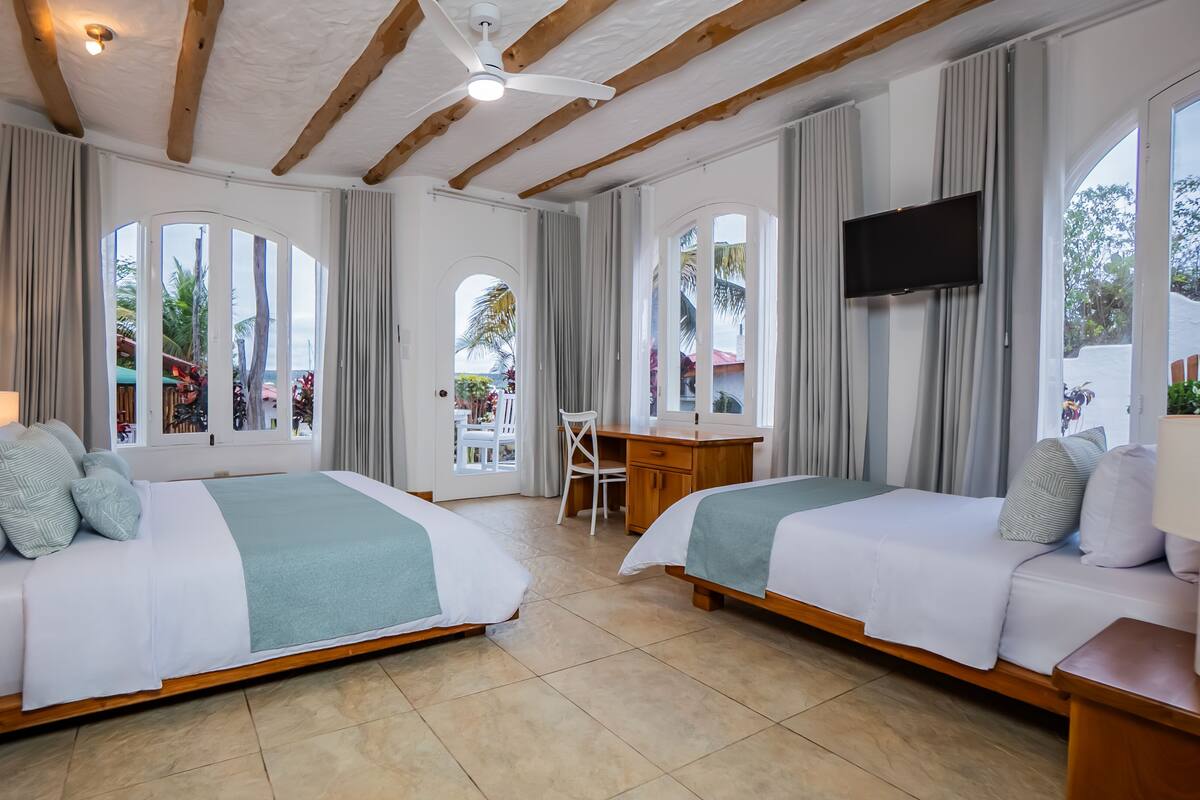 A hotel room with two beds, a desk, a TV, and large windows with curtains.