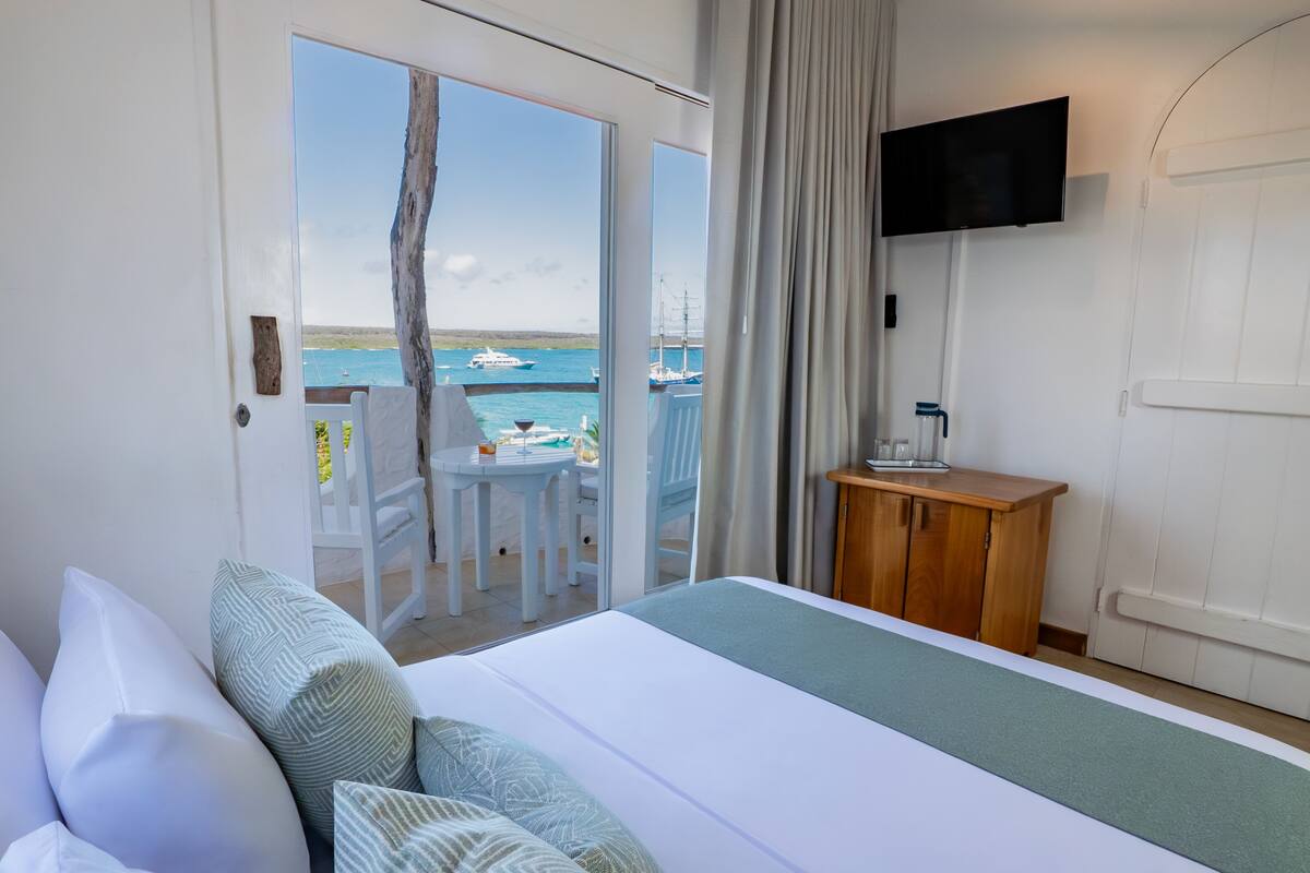 A hotel room with a bed, a TV mounted on the wall, a wooden cabinet, and a balcony with a view of the sea.