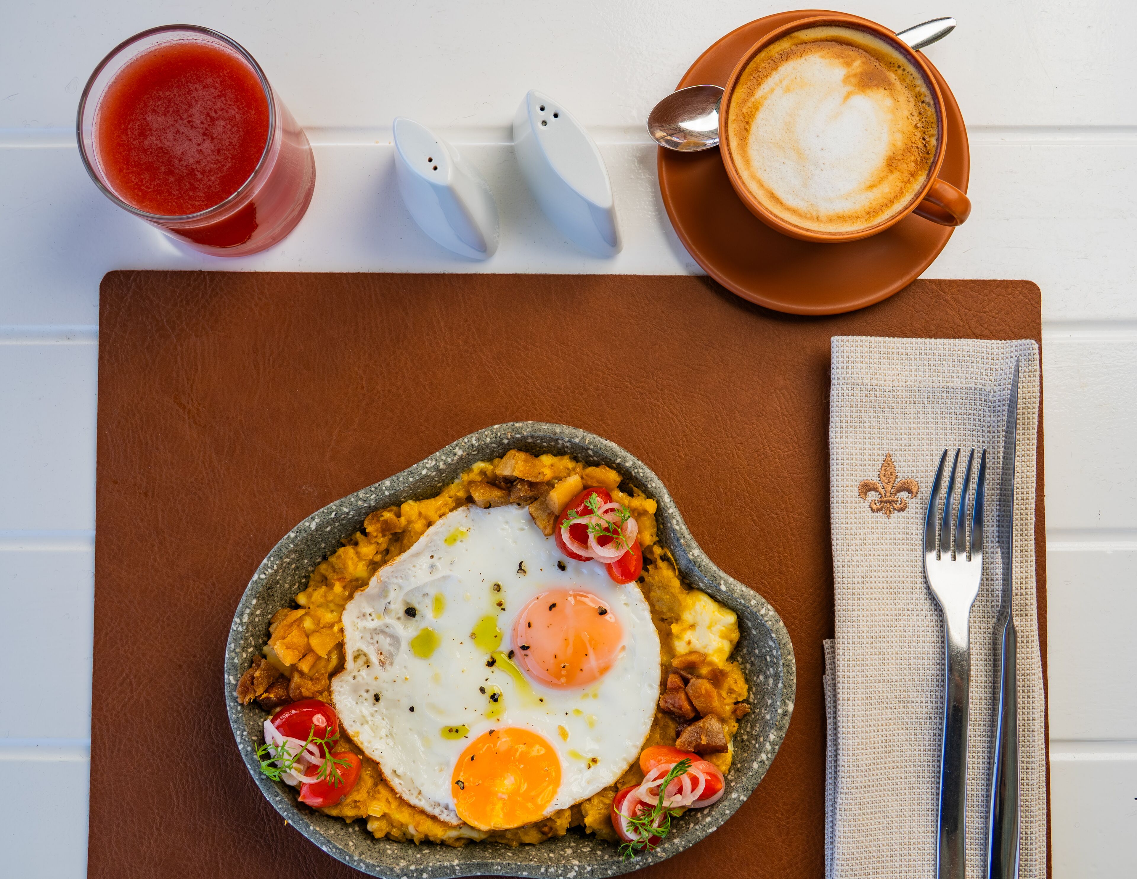 Prato de café da manhã com ovos, tomates e cebolas, uma xícara de café e um copo de bebida vermelha sobre uma mesa.