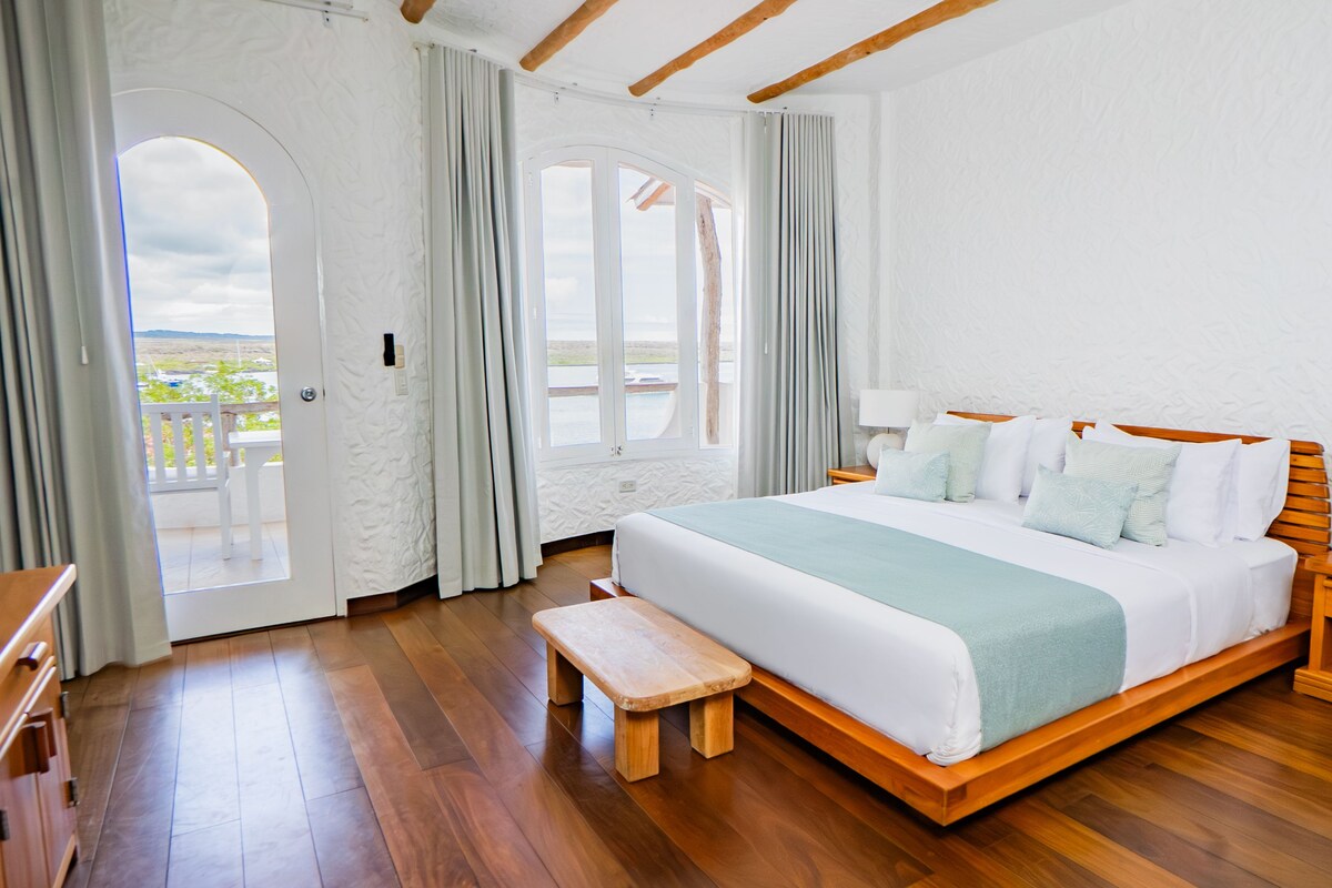 A bedroom with a large bed, wooden floor, a bench, a nightstand, and a view through a door.