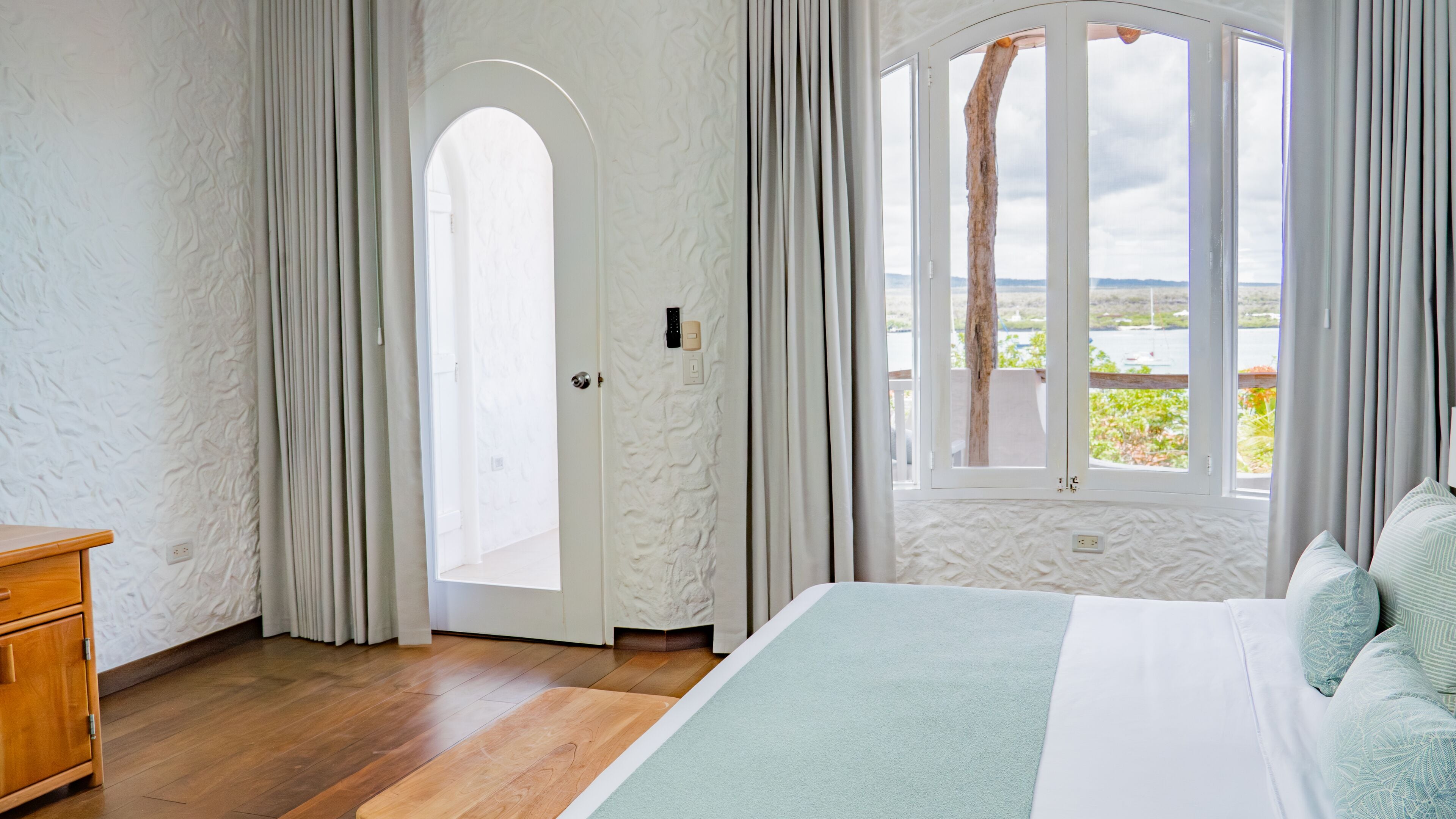 Un dormitorio con piso de madera, una cama con colcha verde y blanca, una mesita de noche de madera, una puerta blanca con ventanal y vistas al exterior.