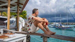 A man sitting on a wooden railing overlooking a marina with boats and a mountainous backdrop.