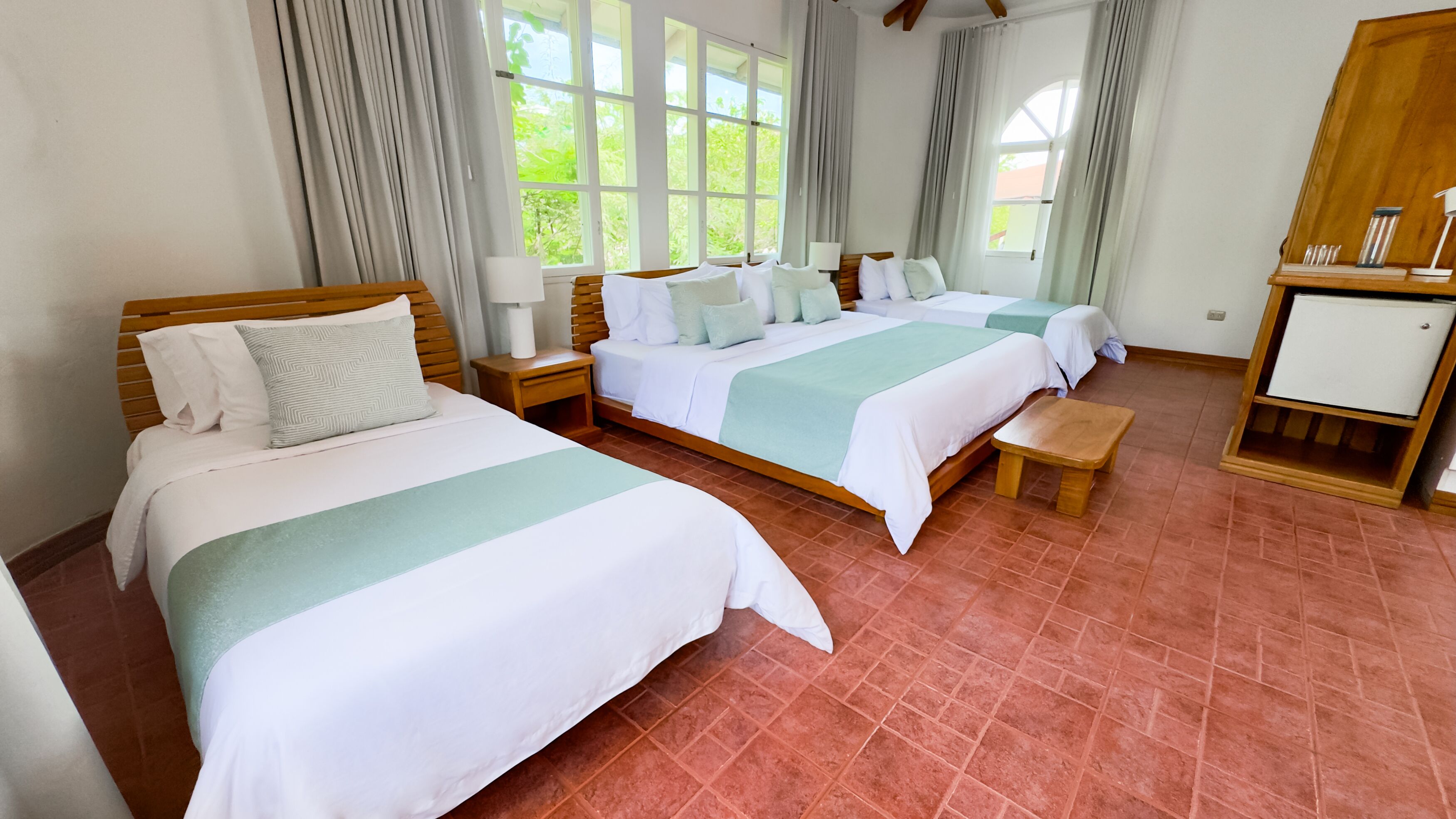 A hotel room with two beds, a wooden bench, a nightstand with a lamp, and a small cabinet.