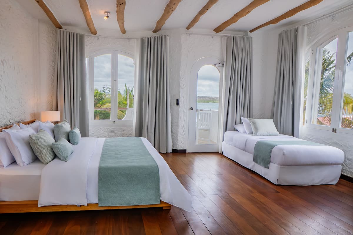 A hotel room with a bed, a sofa, wooden flooring, and large windows with curtains.