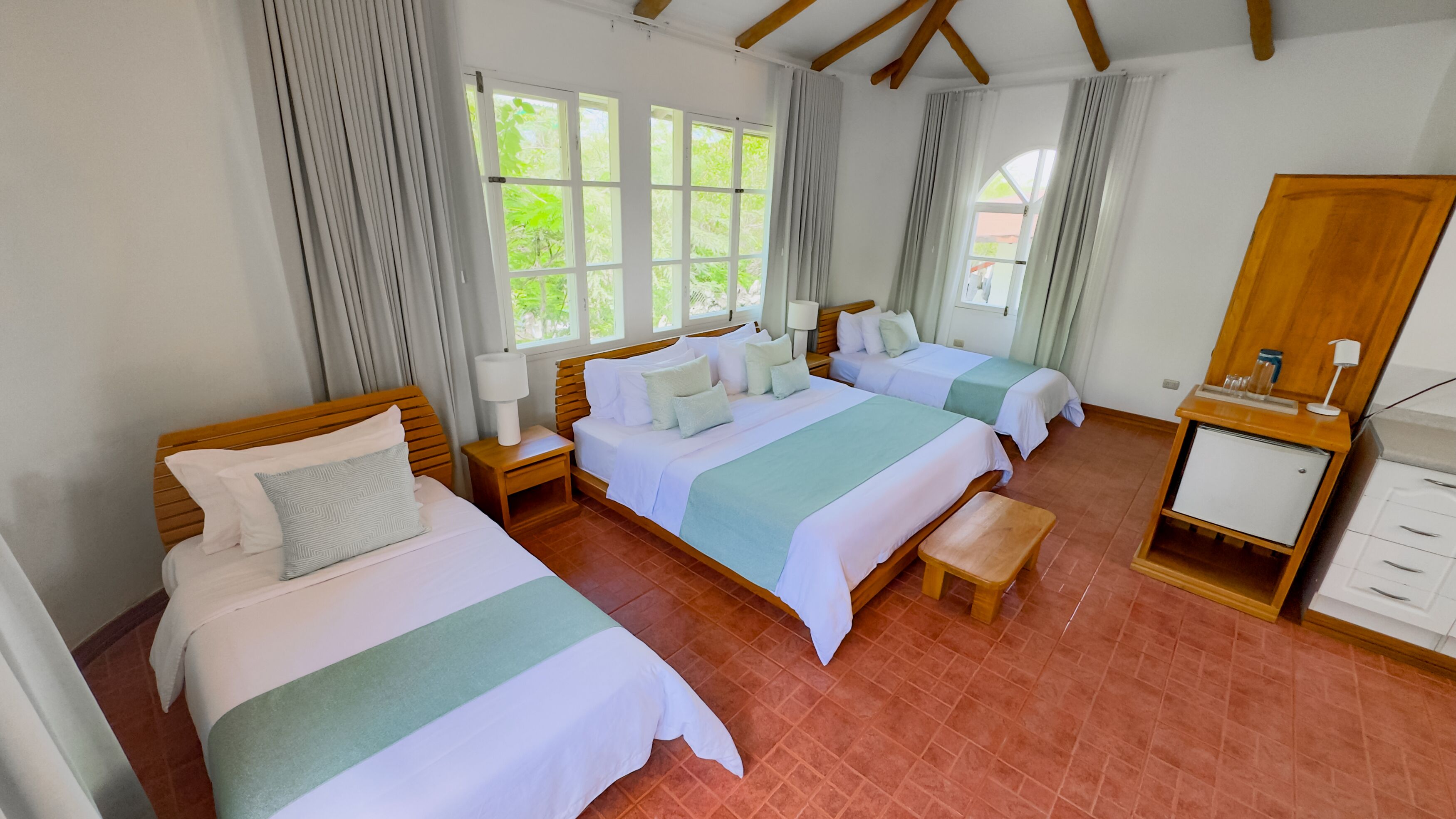 Une chambre d’hôtel avec deux lits, un banc, une table de chevet avec une lampe, une armoire en bois et de grandes fenêtres avec des rideaux.