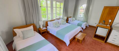 Une chambre d’hôtel avec deux lits, un banc, une table de chevet avec une lampe, une armoire en bois et de grandes fenêtres avec des rideaux.