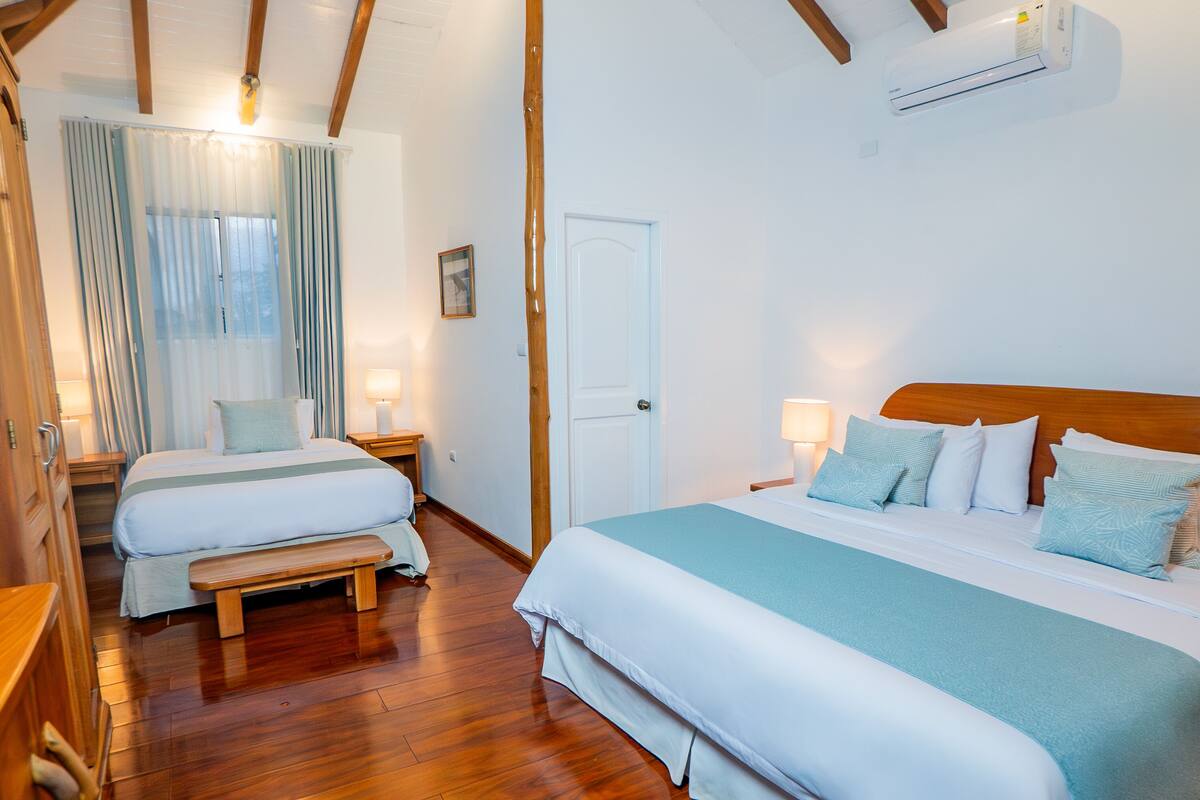 A hotel room with two beds, wooden floors, a wooden headboard, a wall-mounted air conditioner, and a door.
