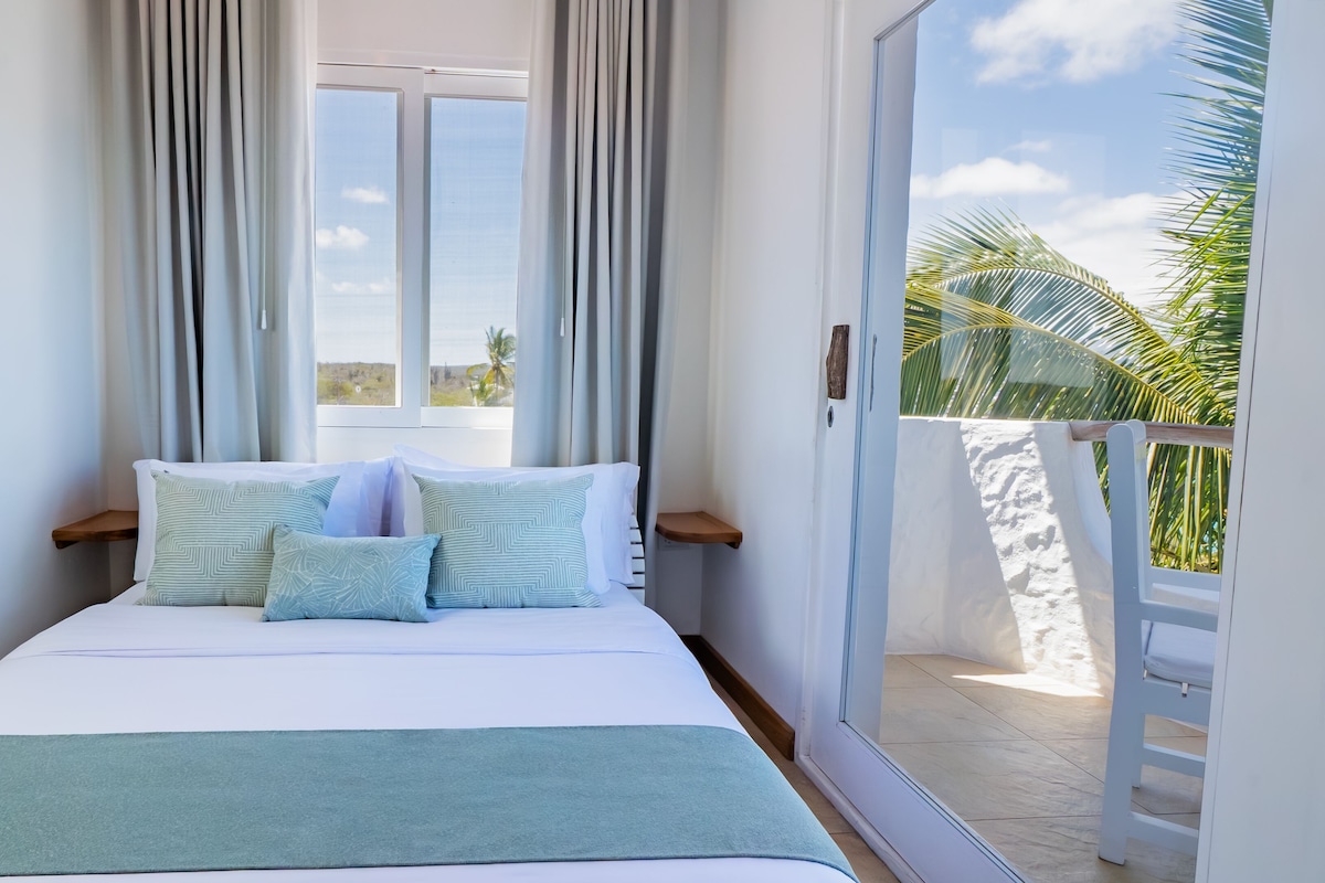 A hotel room with a bed, curtains, a view of palm trees, and a balcony.
