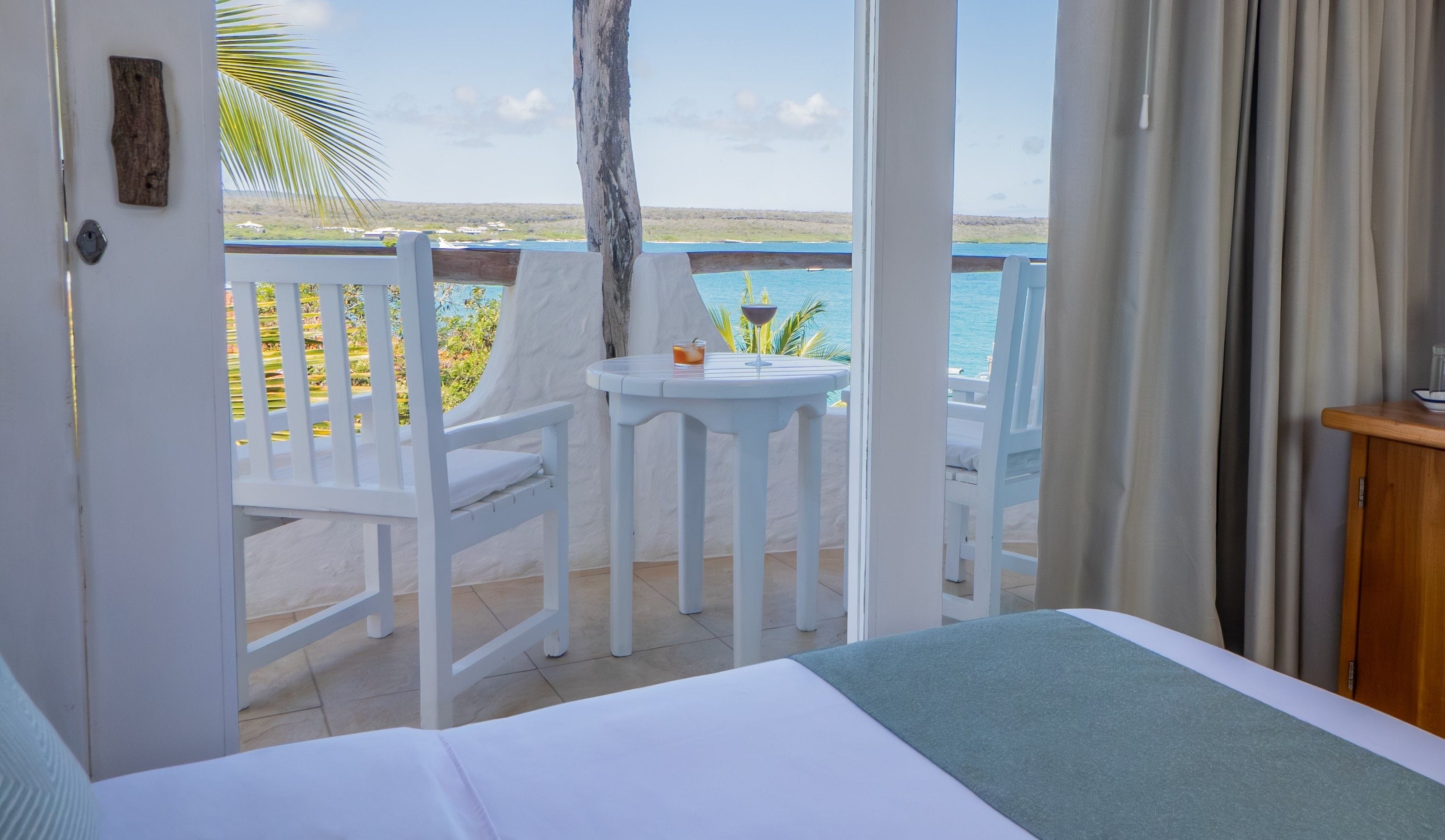 A balcony with a view of the sea, a table with a drink, and a white chair.