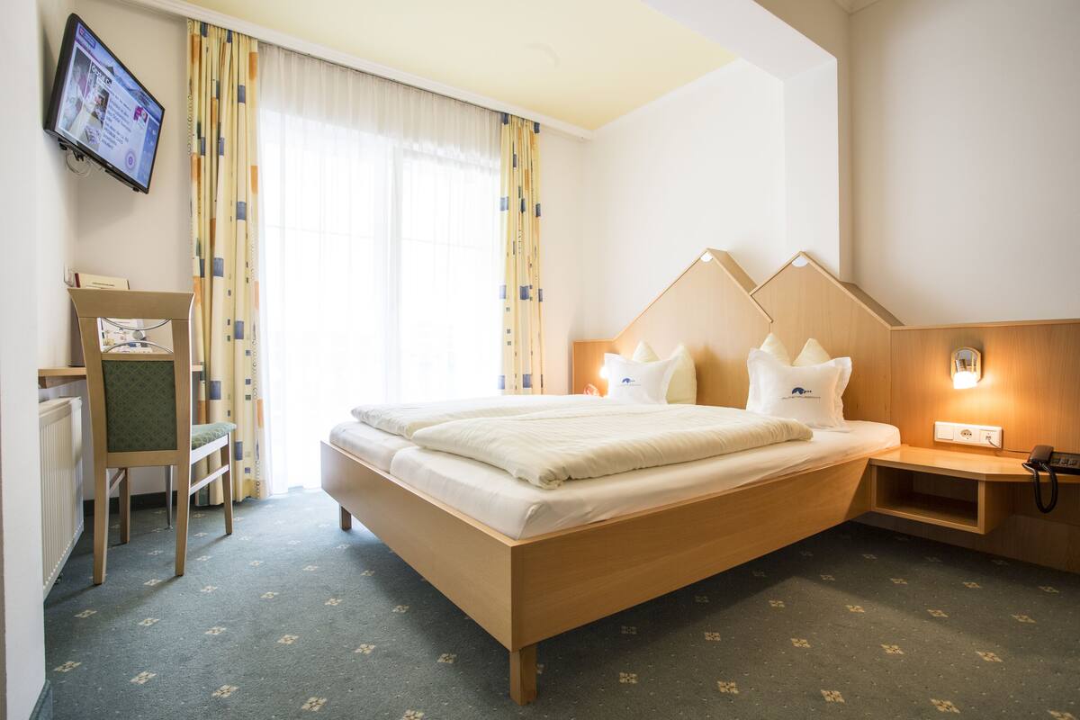 A hotel room with a bed, a desk, a chair, a TV, and a bedside table with a lamp.