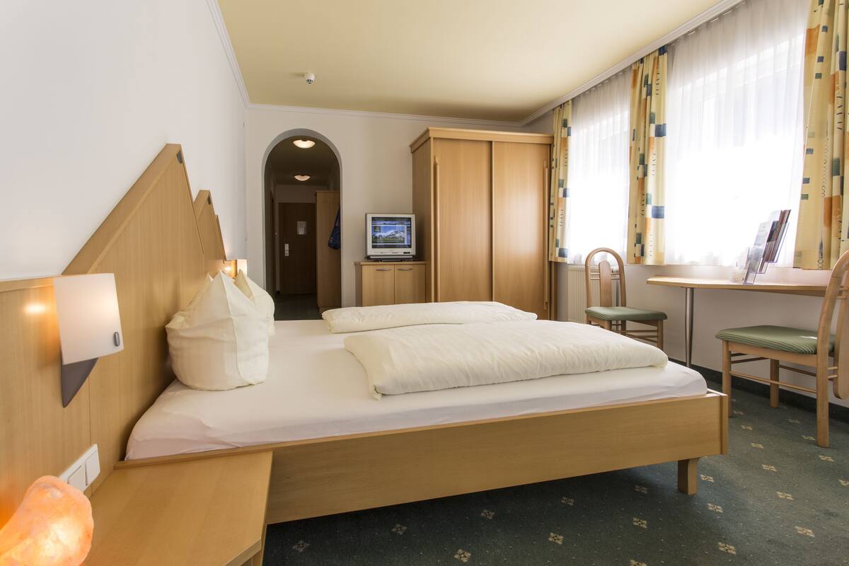 A hotel room with a bed, a desk, a chair, a television, and a bathroom visible in the background.