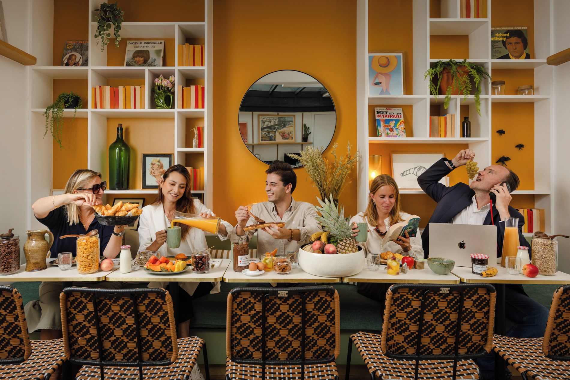 Un groupe de personnes est réuni autour d’une table, où sont disposés divers plats et boissons. Des étagères à livres et des objets décoratifs les entourent.