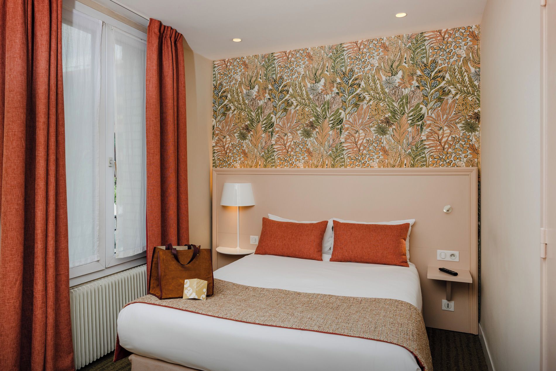 A hotel room with a bed, a nightstand, a lamp, a window with curtains, and a wall with a floral pattern.